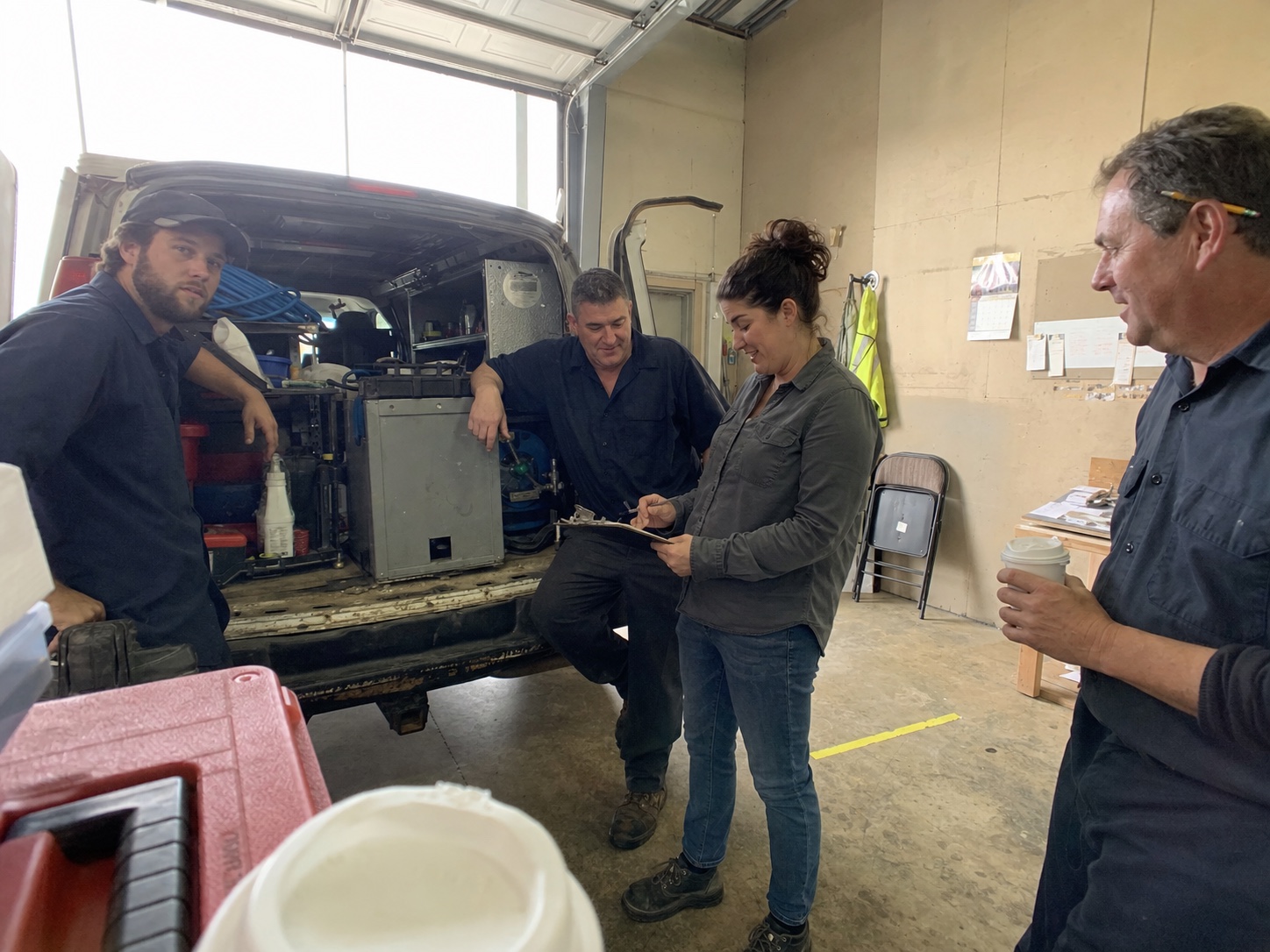 The Lakeland Pest Pros team in the garage at the start of a workday — two field techs and the dispatcher gathered around the open back of the service van, going over the day's stops on a clipboard. Office-manager standing nearby with coffee