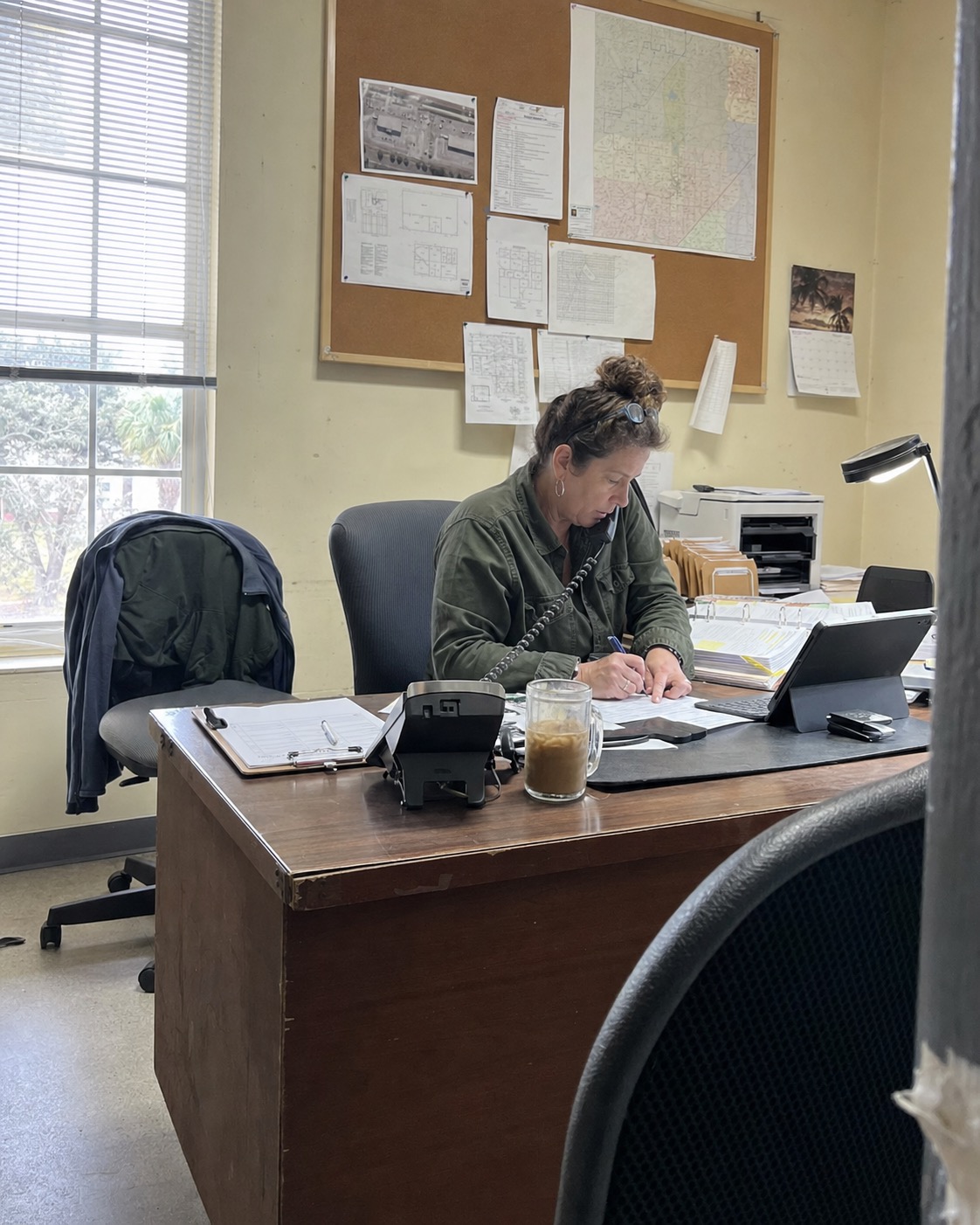 The Lakeland Pest Pros dispatcher mid-call at her desk in the small downtown office — phone wedged between shoulder and ear, pen in hand writing on paperwork. Corkboard behind with pinned Polk County floor plans and a regional map. Window light, sabal palm just barely visible outside